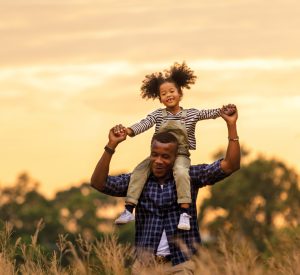man with little girl sitting on his shoulders and holding her hands with an orange sky and trees in the background